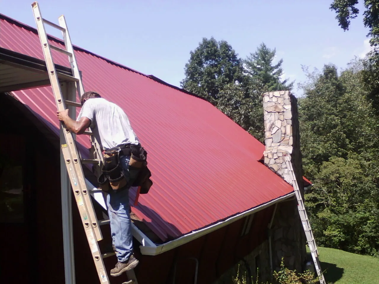 Roofer on a ladder beside a red metal roof and stone chimney — Soffit & Fascia Repair by Roofing Brothers in Haltom City